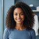 Smiling young woman with dark curly hair and warm brown skin, wearing a slate blue top, against a soft dusty blue background.